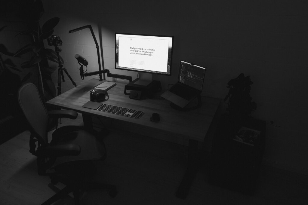 A black and white photo of Statamic web developer Johannes Schuba's desk. You can see a large screen with his website open, a laptop, a mouse, and a keyboard on the table. In front of the desk is a desk chair, next to it a rolling container with a plant on it and the coffee magazine "Standart".