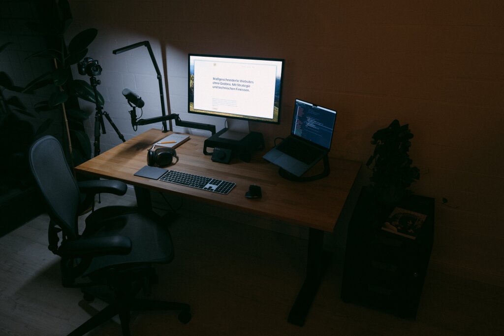 A photo of Statamic web developer Johannes Schuba's desk. You can see a large screen with his website open, a laptop, a mouse, and a keyboard on the table. In front of the desk is a desk chair, next to it a rolling container with a plant on it and the coffee magazine "Standart".