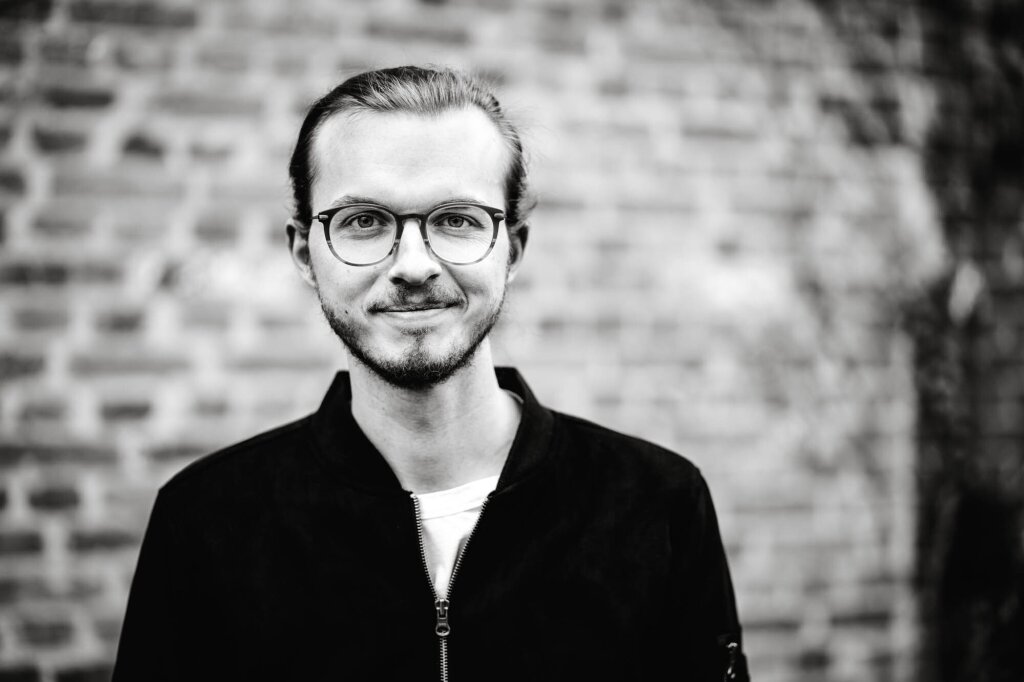 Portrait photo of web developer Johannes Schuba. He wears glasses, smiles lightly into the camera and wears his hair in a bun.