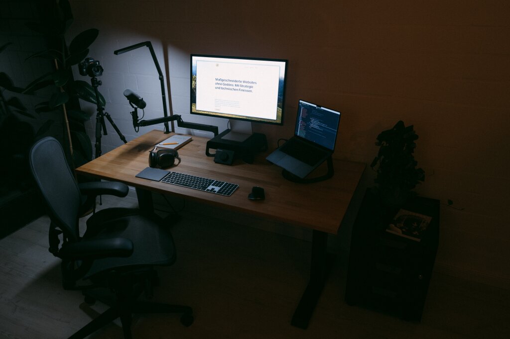 Ein Farbfoto vom Schreibtisch von Statamic-Webentwickler Johannes Schuba. Zu sehen sind ein großer Bildschirm mit seiner Website geöffnet, ein Laptop sowie Maus und Tastatur auf dem Tisch. Vor dem Tisch ein Schreibtischstuhl, daneben ein Rollcontainer mit einer Pflanze darauf sowie dem Kaffee-Magazin „Standart“.